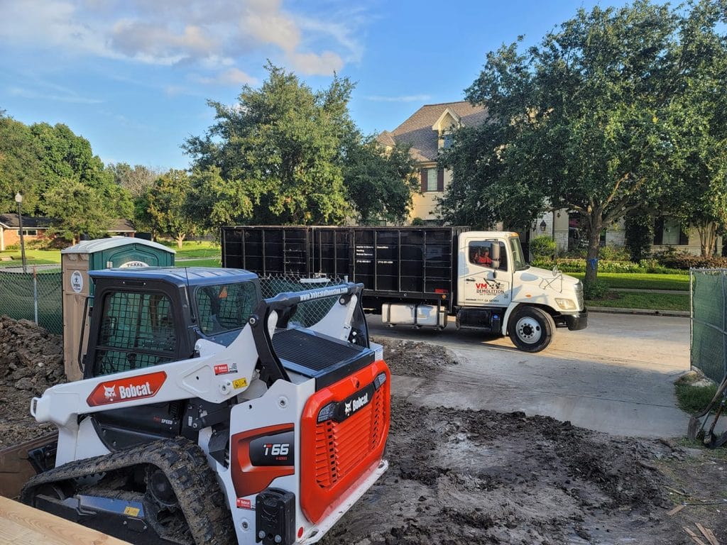 Bobcat T66 on demolition site with VM Demolition truck and portable restroom, surrounded by trees and residential area in Houston, TX.