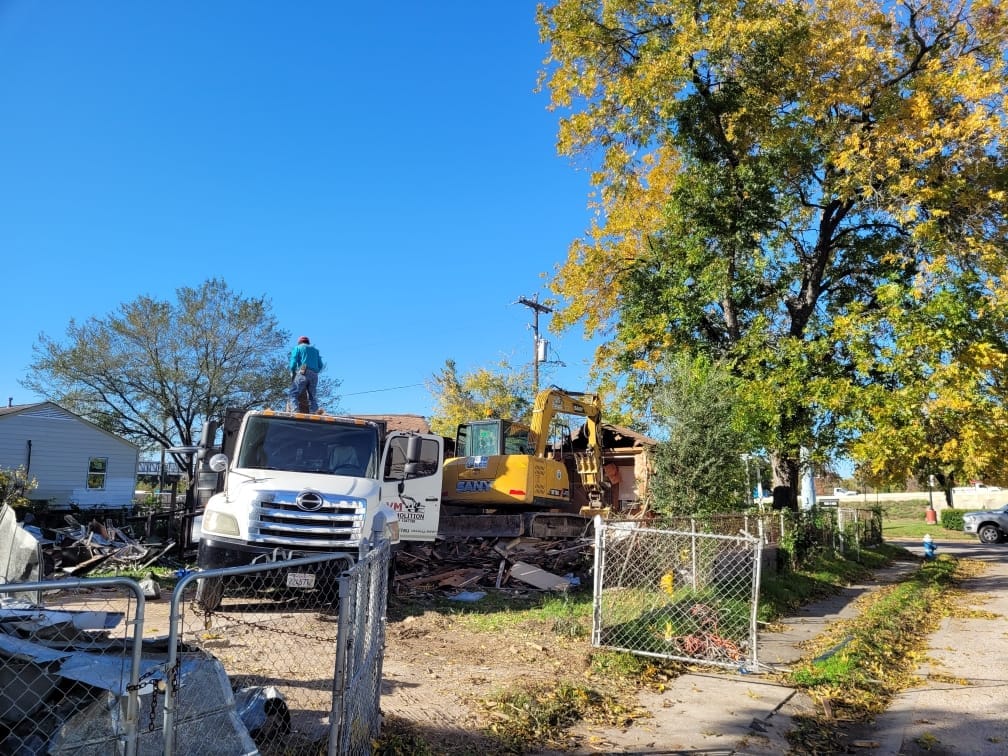 Construction site with VM Demolition truck and excavator, clearing debris from a demolished building, surrounded by trees and residential structures.