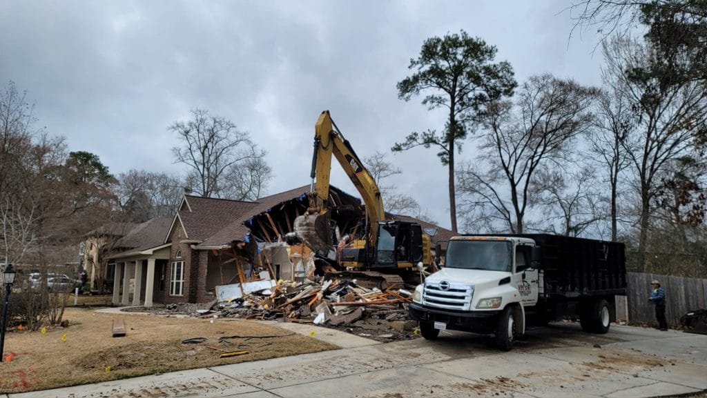 Excavator demolishing a house with debris scattered on the ground, alongside a truck for debris removal, showcasing VM Demolition's construction cleanup services in Houston.