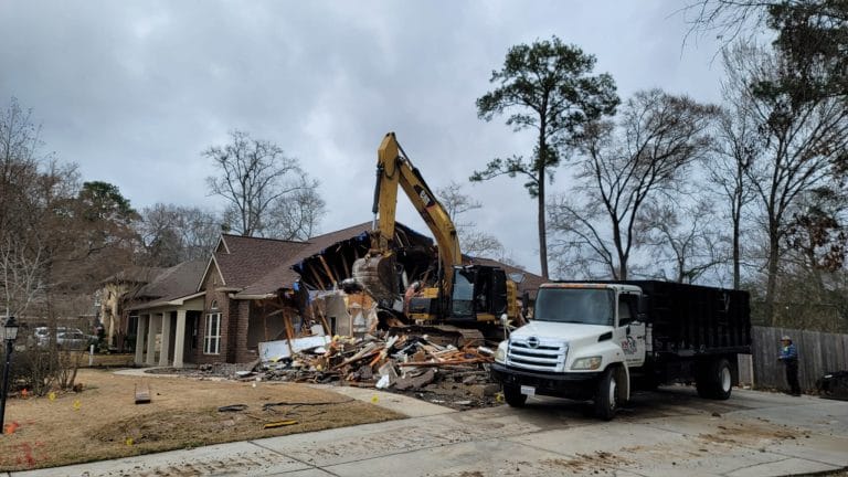 VM Demolition excavator tearing down an older ranch house with a dump truck hauling debris, typical of Clear Lake, Houston teardowns