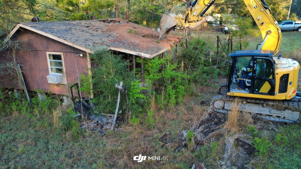 Excavator demolishing an overgrown, dilapidated residential structure in a grassy area, showcasing VM Demolition's expertise in full home demolition services in Houston.